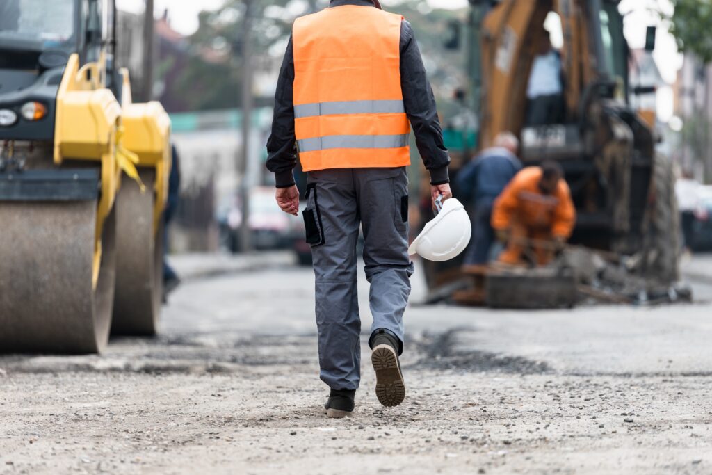 Construction worker wearing safety vest holding hard hat while walking through road work site.