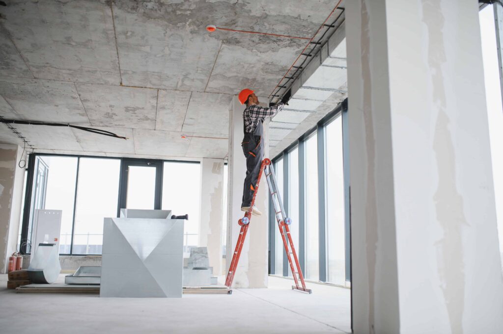 Construction worker standing on ladder installing ceiling wiring inside unfinished building.