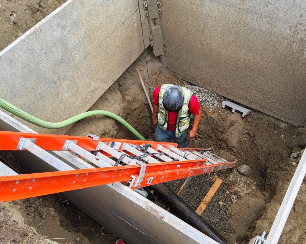 Construction worker in safety vest and hard hat working inside trench with ladder and utility pipes.