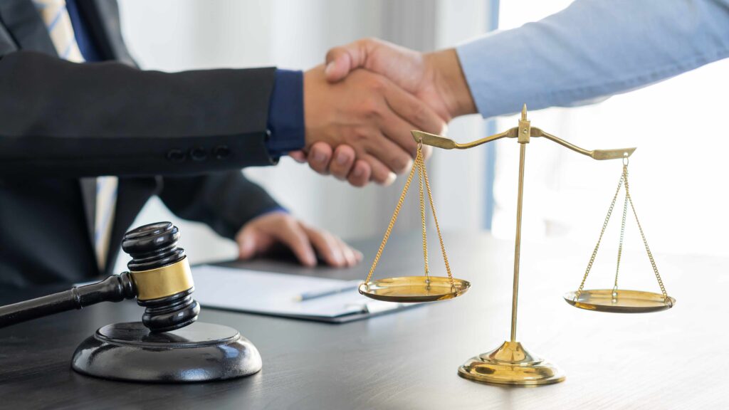 Lawyer and client shaking hands over a desk with a judge&rsquo;s gavel and scales of justice, symbolizing a legal agreement or successful case settlement.