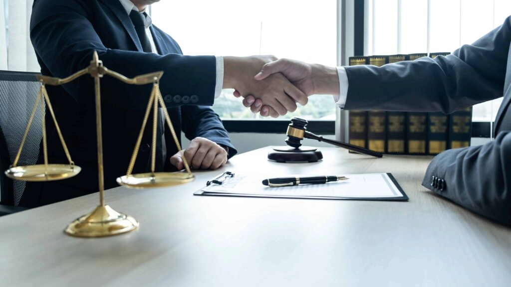 Lawyer and client shaking hands in office with justice scales and gavel on desk.